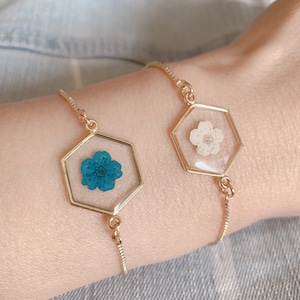 May include: Two gold chain bracelets with hexagonal charms featuring pressed flowers. One charm has a white flower, the other a teal blue flower.