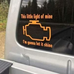 May include: Orange decal on a vehicle window featuring an engine symbol and the text "This little light of mine, I'm gonna let it shine." The decal is likely a humorous take on a vehicle's check engine light.