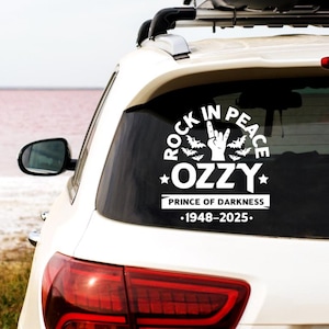 May include: White vinyl car decal featuring the text "ROCK IN PEACE" in a circular design with bats and a hand gesture. Below, the text reads "OZZY" with stars, and "PRINCE OF DARKNESS" with the years 1948-2025.