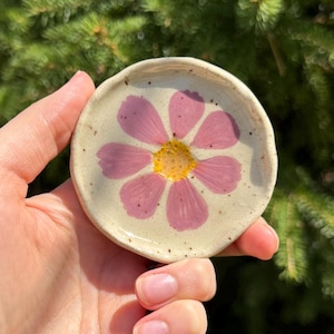 May include: A small, round ceramic dish featuring a hand-painted pink and yellow flower design. The dish has a speckled, off-white background and a raised edge. The flower has pink petals and a yellow centre.