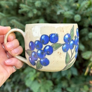 May include: A ceramic mug with a speckled cream-coloured glaze featuring a hand-painted design of clusters of bright blue blueberries on green stems and leaves. The mug has a rounded body and a curved handle on the left side of the vessel.