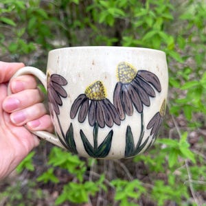 Peut inclure: Une tasse en céramique beige avec une anse, décorée d'illustrations de fleurs marron et jaunes. Les fleurs ont des pétales brun foncé et des centres jaunes. Des feuilles vertes sont peintes à la base des fleurs.