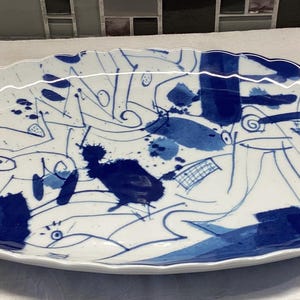 Może przedstawiać: Owalny, biały ceramiczny talerz z falistym brzegiem. Talerz zdobiony jest abstrakcyjnymi niebieskimi pociągnięciami pędzla, plamami i kształtami, tworzącymi nowoczesny, artystyczny wzór.