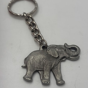 May include: A silver-toned metal keyring featuring a detailed elephant charm attached to a sturdy chain and a circular split ring. The elephant figure displays textured skin details, a raised trunk, and large ears, crafted in a metallic finish.