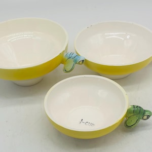 Könnte beinhalten: Drei Keramikschalen mit gelber Außenseite und weißer Innenseite. Jede Schale hat einen einzigartigen Griff mit grünem und blauem Design. Auf der kleinsten Schale steht "1-cup".