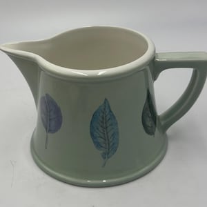 May include: A light sage green ceramic creamer pitcher featuring a smooth, glossy finish and a white interior. The exterior is decorated with three distinct painted leaves in shades of purple, blue, and dark green, positioned around the body.