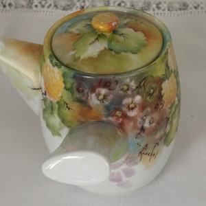 May include: A hand-painted ceramic teapot with a lid. The teapot features a floral design in shades of yellow, green, and purple. The spout and handle are white. The lid has a small, round knob. The teapot is signed.