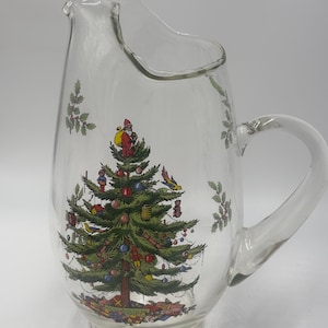 May include: A clear glass pitcher featuring a colourful illustration of a decorated Christmas tree with ornaments, a Santa Claus figure at the top, and wrapped gifts at the base. The design includes green holly sprigs and a curved handle.