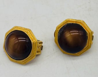 Könnte beinhalten: Ein Paar Vintage-Ohrclips. Jeder Ohrring hat einen großen, runden, braunen Stein, der in einen goldfarbenen, achteckigen Rahmen gefasst ist. Die Ohrringe haben ein klassisches, elegantes Design.