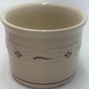 May include: A cream-coloured ceramic crock featuring a cylindrical shape with a wide opening. The exterior is decorated with a subtle embossed brick-like pattern and small, dark maroon floral accents and wavy lines encircling the middle section.