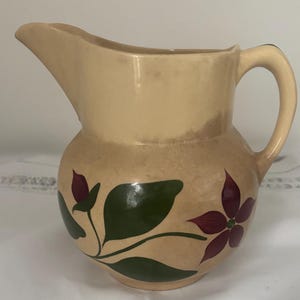 Può includere: Brocca in ceramica beige con corpo arrotondato e manico curvo. La brocca presenta un motivo floreale con foglie verdi e fiori bordeaux. Il beccuccio è curvo per versare facilmente. Un oggetto decorativo per la cucina.