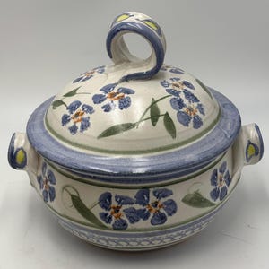 May include: A ceramic lidded bowl with two side handles and a loop handle on the lid. The off-white surface features hand-painted blue flowers with orange centers, green leaves, and blue bands around the rim and base with a small cross-hatch pattern.