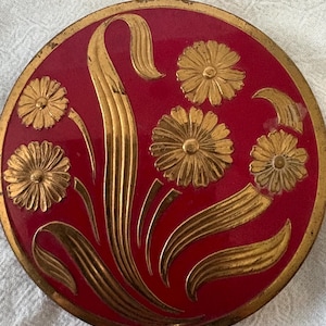 May include: A round, flat metal compact with a deep red enamel finish. The surface features embossed gold-toned floral designs, including four daisy-like flowers and long, curved leaves. The outer edge of the circular lid has a gold-toned rim.