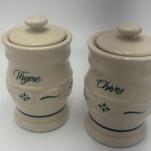 May include: Two off-white ceramic canisters with matching lids. Each canister features a textured basket-weave pattern around the base and a dark green script label reading Thyme on one and Chives on the other, with a small floral accent.