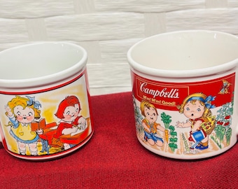 May include: Two white ceramic mugs with a red band around the top and a handle. Each mug features a vintage Campbell's Soup Kids illustration. One mug has the text "Campbell's, M'm! M'm! Good!"