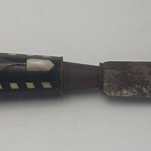 May include: A knife with a dark, weathered metal blade and a rectangular handle. The handle is black with a white checkered pattern along the edges, featuring decorative metal inlays and a small white stone accent on the top surface.