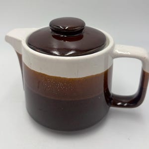 Puede incluir: Una tetera de cerámica con un diseño de dos tonos, con una base de color marrón oscuro brillante y una sección superior blanca. Incluye una tapa a juego de color marrón oscuro con un pomo central, un pico corto y un asa cuadrada lateral.