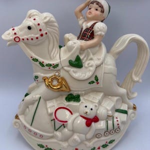 May include: A ceramic teapot shaped like a rocking horse with a child rider. The horse is cream-coloured with red dots, green holly leaves, and a gold horn. A white teddy bear with a red bow sits at the base among gift boxes. The child wears a plaid vest.