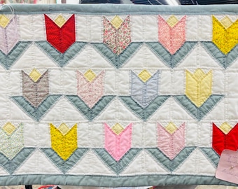May include: A handmade quilt with a repeating tulip pattern. The tulips are in various colors, including red, yellow, pink, and floral prints, set against a white background with a light blue border. The quilt is approximately 24 inches wide and 36 inches long.