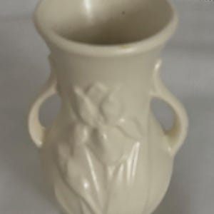 Puede incluir: Jarrón de cerámica color crema con dos asas pequeñas y un diseño floral en relieve. El jarrón tiene un cuerpo bulboso y un borde acampanado. Una pieza decorativa para exhibir flores o como acento independiente.