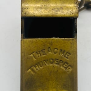 Puede incluir: Primer plano de un silbato de latón antiguo. El texto "THE ACME THUNDERER" está grabado en relieve en la parte delantera. El silbato tiene un lazo para un cordón y muestra signos de edad y uso.