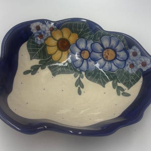 Könnte beinhalten: Eine blattförmige Keramikschale mit tiefblauem Rand und cremefarbener Mitte, die mit kleinen dunklen Punkten versehen ist. Das Innere ist mit einem handgemalten Blumenmotiv verziert, das eine gelbe Sonnenblume, zwei blaue Blüten und kleine blaue Blumen zeigt.