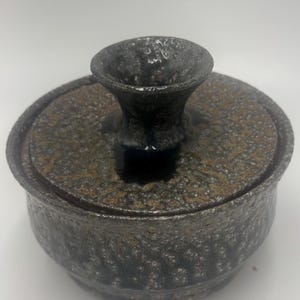 May include: A round ceramic lidded bowl with a textured, mottled surface in shades of dark gray, black, and metallic bronze. The lid features a prominent, flared, funnel-shaped handle with a rough, dark finish that contrasts with the bowl.