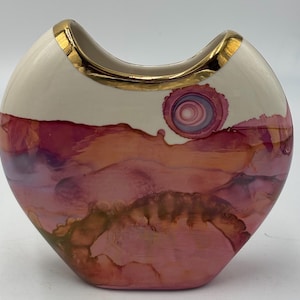 May include: A crescent-shaped ceramic vase featuring a smooth cream-colored upper section and a vibrant lower portion decorated with swirling shades of pink, magenta, and soft orange ink-like patterns, finished with a metallic gold rim.