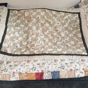 Puede incluir: Un edredón hecho a mano con un diseño de patchwork. El centro presenta un patrón de cuadros en beige y blanco, enmarcado por un borde marrón oscuro. Los bordes muestran patrones florales y geométricos en varios colores.