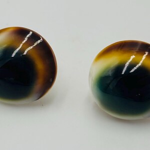 May include: Two polished, round cabochons displaying a gradient of colours. The stones transition from a dark, almost black centre, through shades of green, yellow, and brown, to a creamy white edge. The polished surface reflects light.