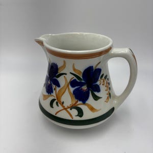 May include: White ceramic pitcher with a handle and spout. The pitcher is decorated with blue floral designs, brown accents, and a green base. The pitcher is likely used for serving beverages.