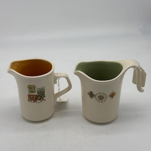 Peut inclure: Deux crémiers en céramique avec des motifs différents. L'un a un bord vert et un motif floral, l'autre a un bord orange et un motif de maison et de chariot.