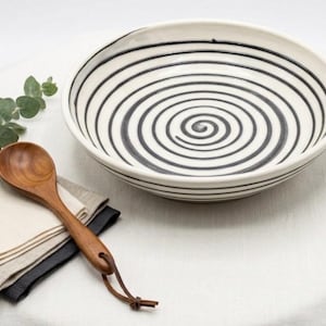 May include: A wide, shallow ceramic bowl featuring a hand-painted black spiral pattern on a cream-colored background. Beside the bowl lies a polished wooden spoon with a leather cord attached to the handle, resting on folded linen napkins.