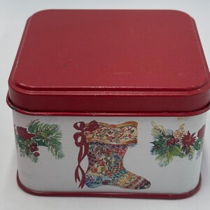 May include: A square metal tin with a solid red lid and a white base. The sides of the tin feature a repeating pattern of a decorative Christmas stocking with a red bow and green pine branches with red berries printed on the white background.