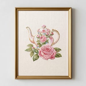 Könnte beinhalten: Gerahmte Stickerei mit einer rosa Teekanne mit Rosen und grünen Blättern. Die Teekanne hat ein Blumendesign mit blauen Akzenten. Unten befindet sich eine große rosa Rose. Der Hintergrund ist ein hellbeiger Stoff.