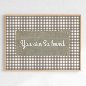 Può includere: Opera d'arte da parete incorniciata con sfondo a motivo gingham marrone e bianco. Una toppa rettangolare in tessuto verde oliva strutturato è centrata, fissata con rivetti in rame e presenta il testo bianco "You are So loved".