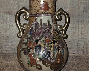 May include: An antique vase with a beige background and gold accents. The vase features a detailed painting of figures in traditional attire, surrounded by flowers. The vase has two gold handles and a flared top.