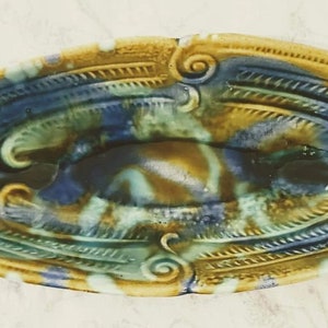 May include: A ceramic dish with an oval shape and a blue, green, and gold glaze. The dish has a raised design around the edge and a smooth interior.