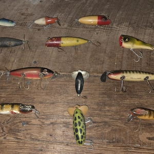 May include: A collection of vintage fishing lures in various shapes and colors, including red, yellow, and green. Some lures have propellers and multiple hooks. The lures are arranged on a wooden surface.