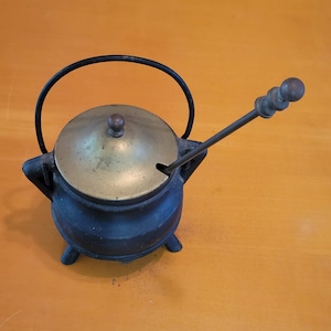 Puede incluir: Una olla de hierro fundido negra con una tapa de latón y un mango largo de madera. La olla tiene un diseño decorativo en el lateral y está sentada sobre una superficie de madera.