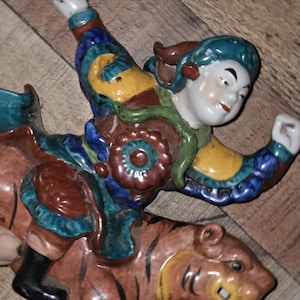Puede incluir: Figura de cerámica que representa a una persona montada en un tigre. La figura viste ropa colorida con detalles azules, amarillos y rojos. El tigre es marrón con rayas negras y una cara amarilla. La figura es probablemente un objeto decorativo.