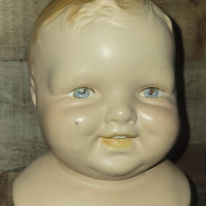 May include: A vintage porcelain doll head with a smiling expression. The doll has blue eyes and a rosy complexion.