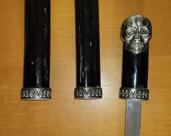 May include: Three black handled daggers with silver skull accents. The center dagger has a silver skull pommel and a silver skull design on the handle. The other two daggers have silver skull designs on the handles.