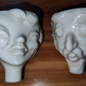 Può includere: Due figurine in ceramica bianca di volti, una con un'espressione più femminile e l'altra con un'espressione più maschile.