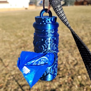 May include: A blue metal dog waste bag dispenser with a decorative design. The dispenser is attached to a black strap.