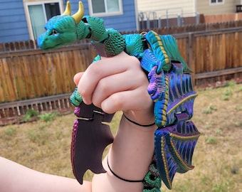 May include: A green, blue, and purple iridescent dragon puppet with gold accents. The dragon is articulated and has wings that can be moved.