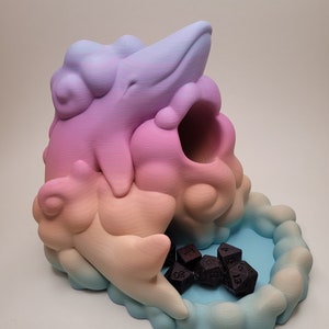 May include: A pastel pink, orange, and blue ceramic dice tower shaped like a cloud with a whale on top. The tower has a small tray at the bottom for holding dice. There are several black dice in the tray.