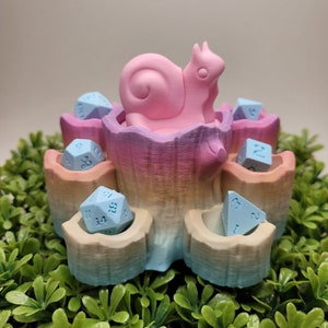 May include: A pastel pink and blue dice tower shaped like a tree stump with a pink snail on top. The tower has six slots for dice, each with a different color gradient. There are six blue dice in the slots.