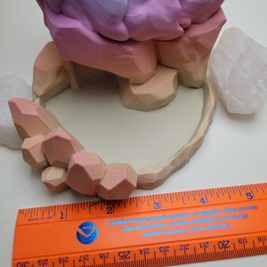 May include: A 3D printed planter shaped like a mountain range with a pink, purple, and orange gradient. The planter is sitting on a white surface with two white crystal rocks. A ruler is placed below the planter to show the size.