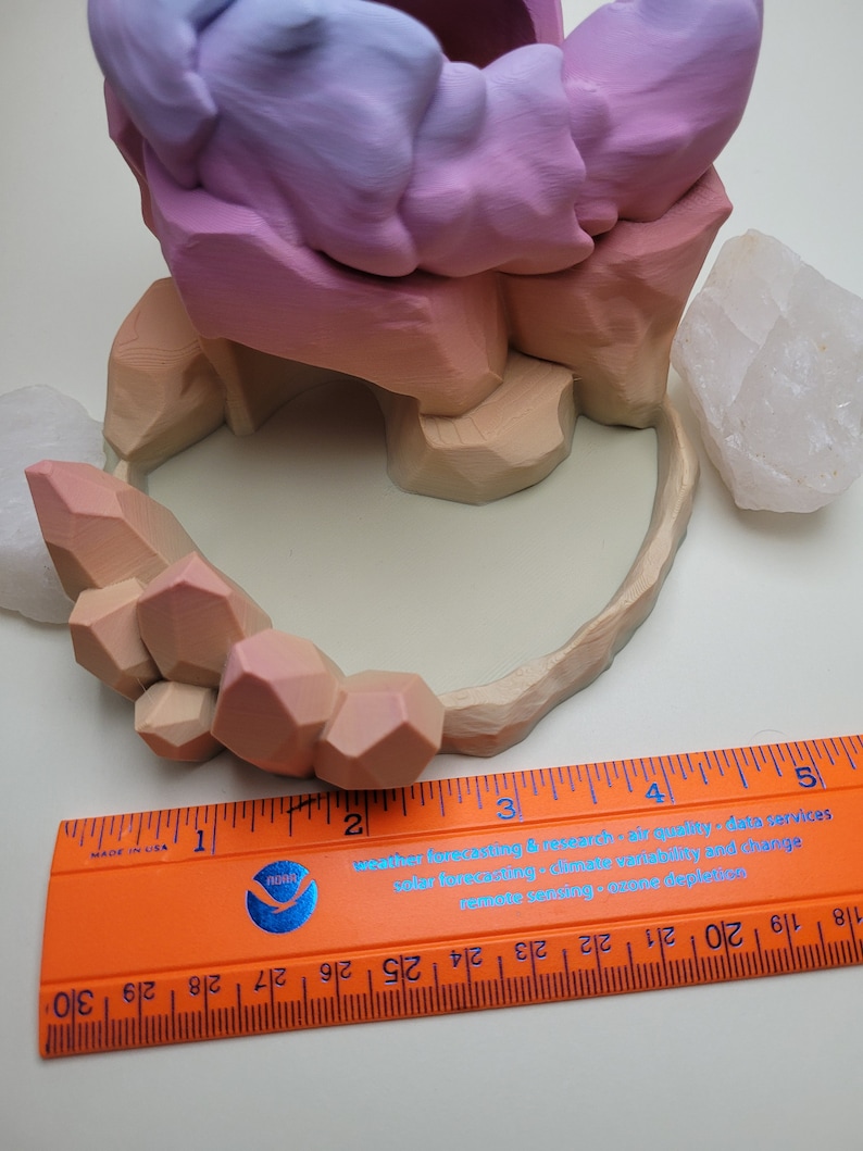 May include: A 3D printed planter shaped like a mountain range with a pink, purple, and orange gradient. The planter is sitting on a white surface with two white crystal rocks. A ruler is placed below the planter to show the size.